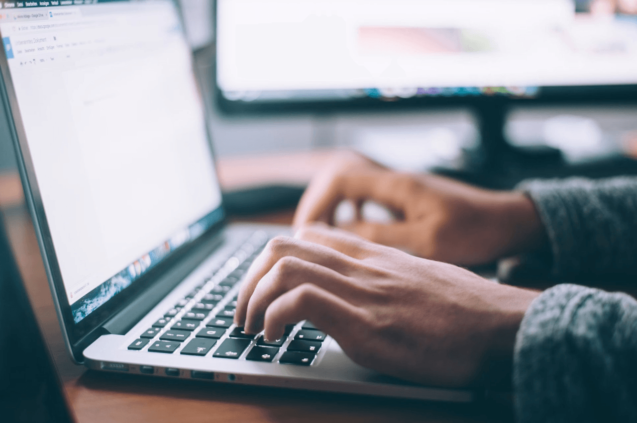 Hands typing on a laptop at a desk with a notebook, glasses, and phone nearby